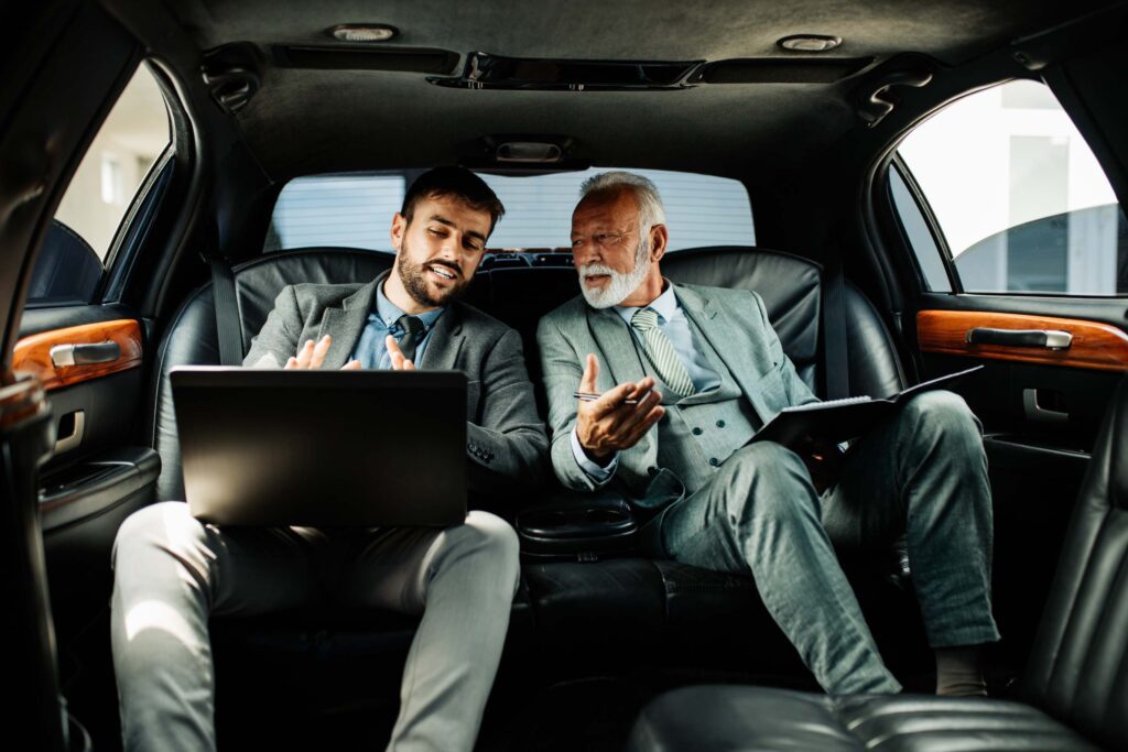 Two men in suits sitting in a car with one of them looking at the other.